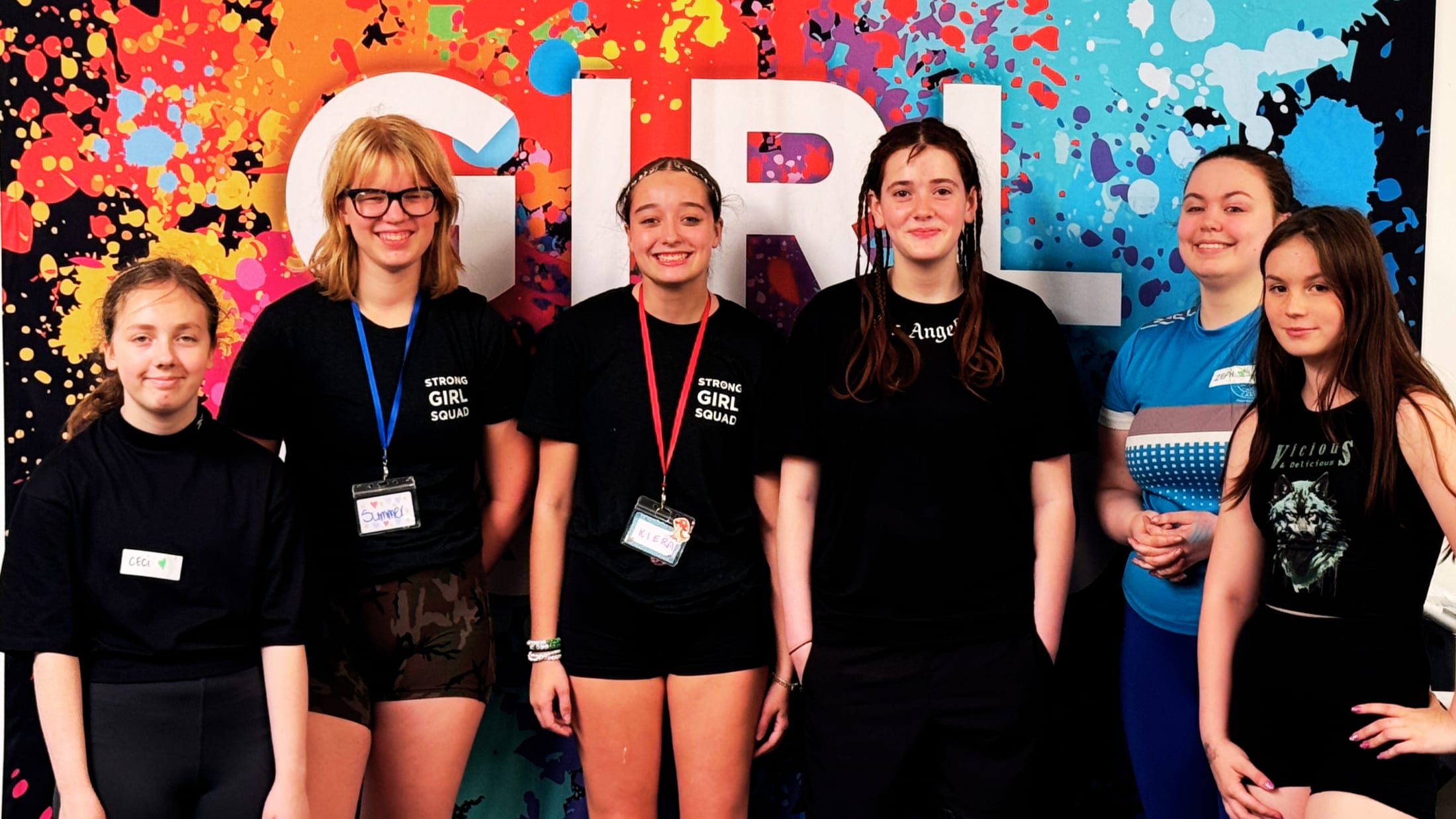 A squad of teenage girls in Strong Girl Squad shirts in front of a colourful GIRL mural