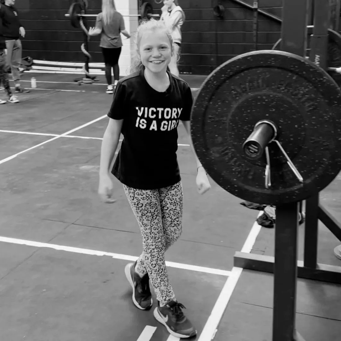 A grinning girl in a ‘Victory is a Girl’ t-shirt by a barbell